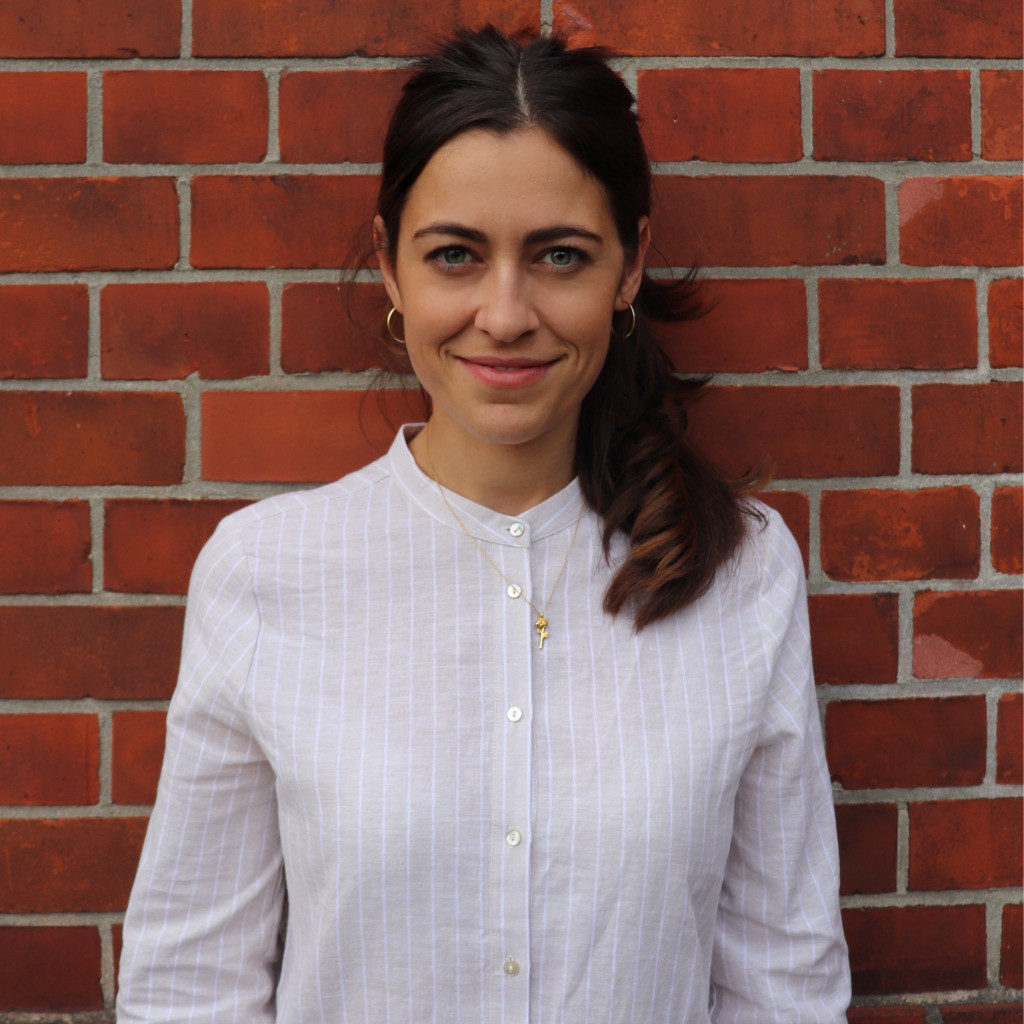 Sarah Schnorrbusch steht lächelnd in weißer Bluse vor einer roten Backsteinwand.